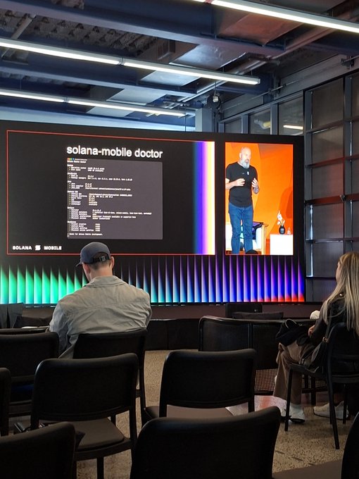 Two people sit in a conference room watching a presentation on a large screen displaying code titled 'solana-mobile doctor' alongside a speaker. The screen shows the Solana logo at the bottom left.