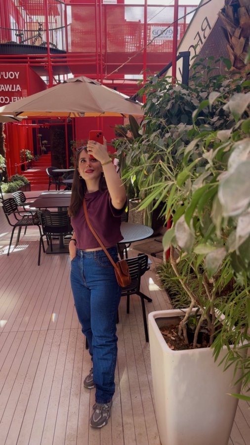 A young woman wearing a maroon top, blue jeans, and a brown crossbody bag takes a selfie on her phone in an outdoor cafe area with red architecture and green plants.
