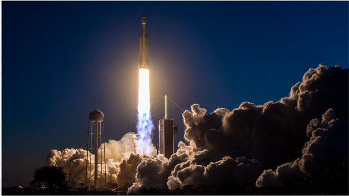 A rocket launches into a clear blue sky, with bright flames and billowing white smoke emerging from the launch pad below.