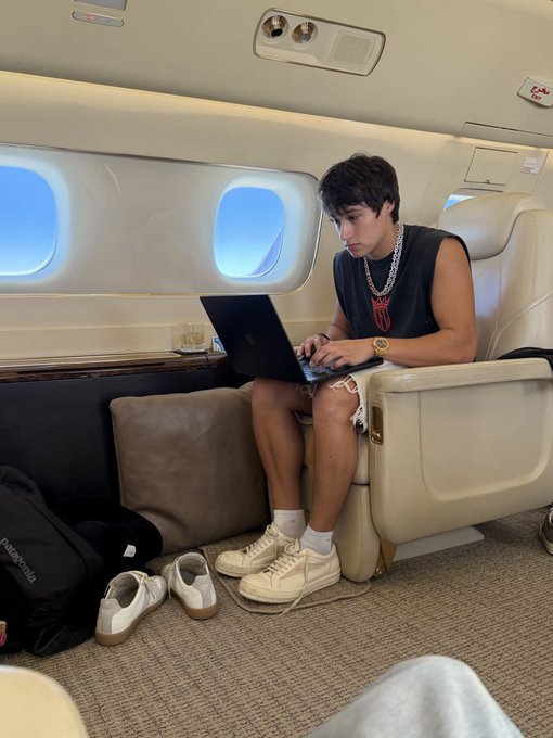 A young man wearing a black tank top, pearl chain, and ripped shorts sits working intently on a laptop inside a private jet, with a Patagonia bag and sneakers on the floor beside him. The image visually supports the post's joke about struggling to be productive, suggesting that even a luxurious setting like a private jet isn't enough to make the work happen.