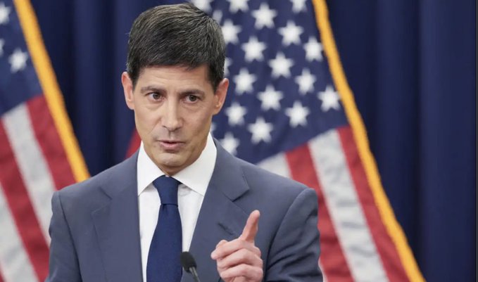A man in a dark suit and blue tie speaks at a podium, gesturing with his index finger, with American flags displayed in the background.
