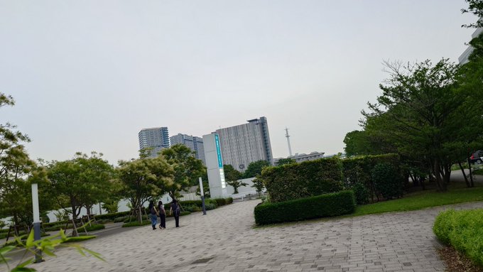 An outdoor park scene with a paved walkway, green trees, hedges, and people walking, with high-rise buildings visible in the background under an overcast sky.