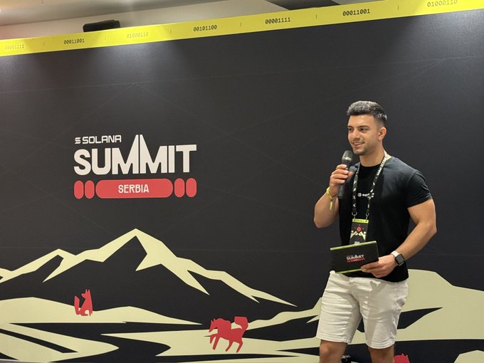 A man speaks into a microphone at the 'Solana Summit Serbia' event, standing in front of a backdrop with mountain graphics and binary code pattern tape.