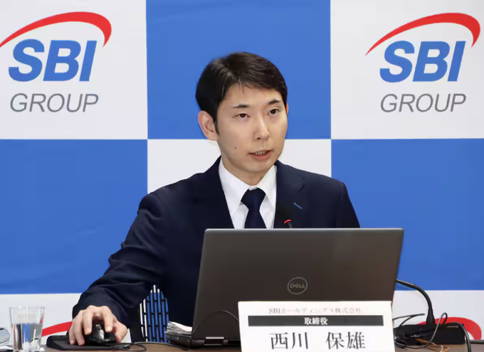 A man in a dark suit speaks at a press conference with an "SBI Group" logo backdrop, seated behind a laptop and a nameplate reading "西川 保雄" (Yasuo Nishikawa) of SBI Holdings, as referenced in the post about SBI's earnings briefing discussing XRP's current standing and its approximately ¥6.6 trillion value tied to Ripple's holdings.