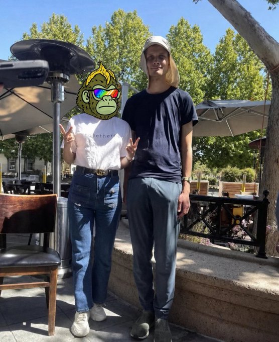 An outdoor photo of two people at a restaurant patio, with one person's face obscured by a colorful Bored Ape NFT emoji sticker, while the other wears a black t-shirt and cap.