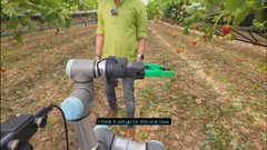 A robotic arm holding a green spray bottle outdoors near plants, with a person in a yellow shirt standing in the background.