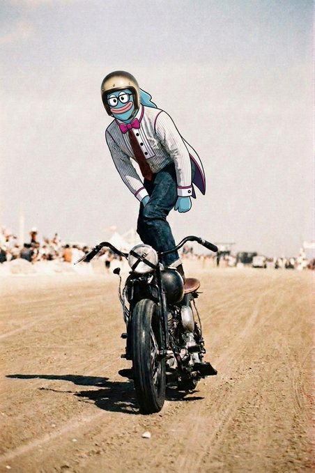A person wearing a helmet, goggles, and a clown mask with a pink bow tie and suspenders stands on the seat of a motorcycle in a desert landscape, giving a thumbs-up.
