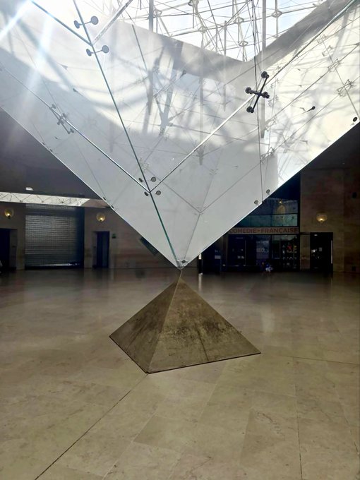 An inverted glass pyramid is suspended above its smaller stone counterpart in the Louvre's lobby, with "LIBRAIRIE FRANÇAISE" visible in the background. The striking juxtaposition of glass and stone, structure and reflection, visually echoes the digital duality of Bitcoin as both intangible code and tangible value.