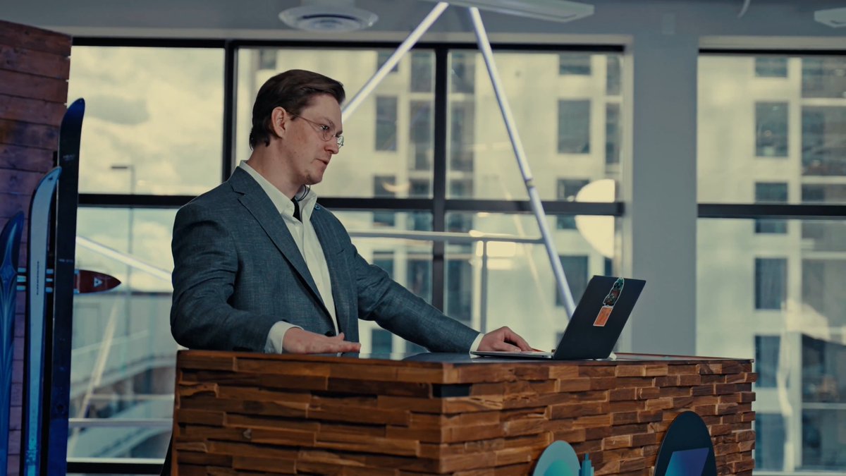 A man in a gray suit and glasses stands behind a wooden podium, focused on a laptop with a lanyard, while skis are propped against the wall in a modern room with large windows overlooking city buildings. The setting suggests a presenter preparing for the MtnDAO Demo Day live stream announcement.