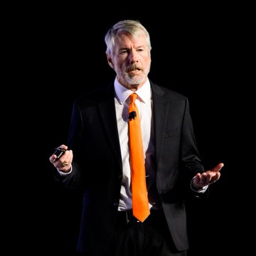 urn of orange liquid, a black object with a white face and a black background, and a man in a suit speaking at a podium against a black backdrop.