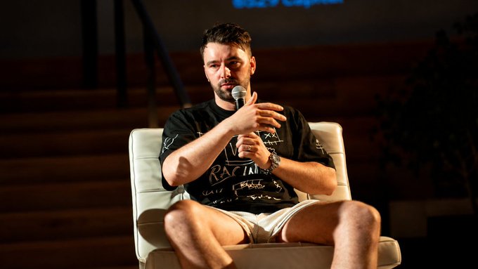 A bearded man sits on a white chair holding a microphone, wearing a black graphic t-shirt and light shorts on what appears to be a stage.