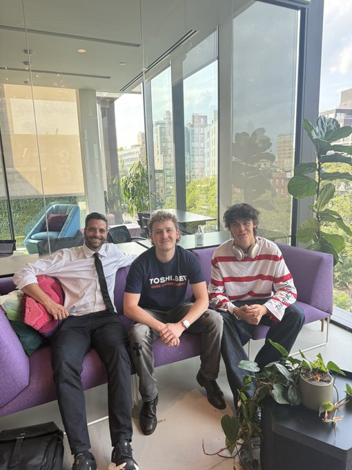 Three young men sit smiling on a purple couch in a modern office with large windows overlooking a city skyline.