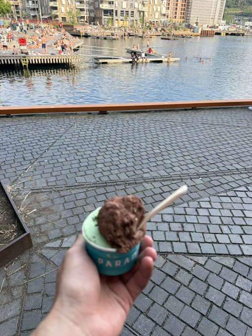 A hand holding a cup of chocolate ice cream with a spoon, overlooking a waterfront with floating docks and people swimming.