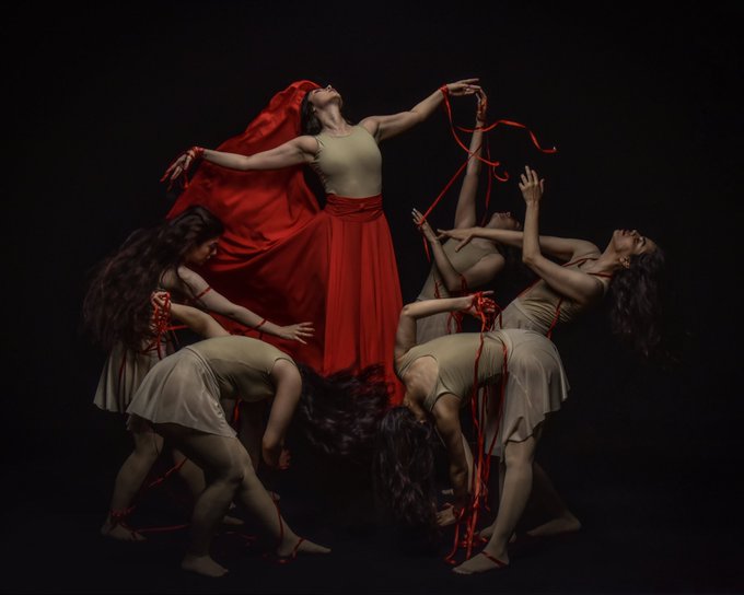 A central dancer in a flowing red skirt and beige top stands elevated, head thrown back and arms outstretched with trailing red ribbons, surrounded by four other dancers in matching attire who crouch and bend dramatically against a stark black background. The striking composition, with its vibrant red accents and theatrical choreography, embodies the emotional intensity and artistic expression characteristic of high-value digital art collectibles on Ethereum.