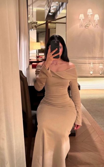Mirror selfie photograph of a woman in an elegant beige off-shoulder form-fitting gown, taken in what appears to be an upscale hotel room with chandeliers and luxury furnishings.