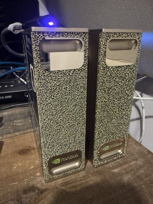 Two textured NVIDIA-branded hardware units (likely GPUs or compute modules) with a speckled green and black finish stand side by side on a wooden surface, with cables and networking equipment visible in the background.