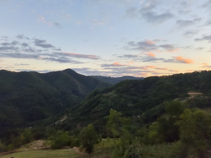 A scenic landscape view of lush green, forested mountains under a sky illuminated by a colorful sunset or sunrise.