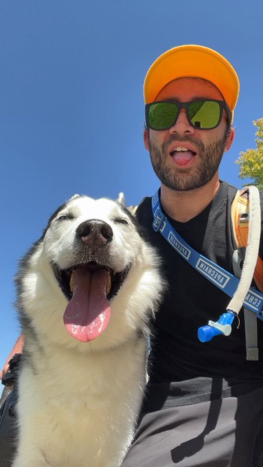 A selfie of a bearded man wearing an orange cap, green reflective sunglasses, and a CamelBak hydration pack posing with a smiling husky dog against a clear blue sky.