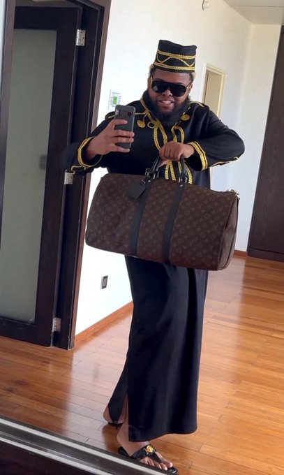 A man wearing sunglasses, a black embroidered cap, and a black and gold embroidered traditional outfit takes a mirror selfie while holding a brown Louis Vuitton monogram duffle bag in a hallway with wooden floors.