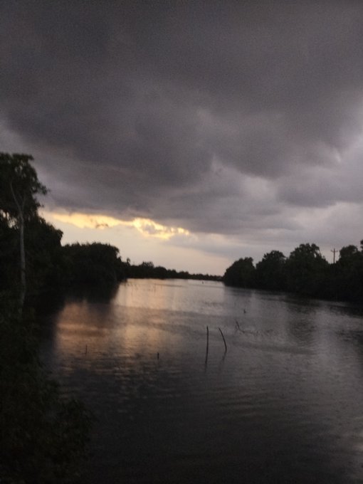 A moody, cloudy sunset over a calm river or lake lined with dark trees, with the light breaking through the storm clouds.