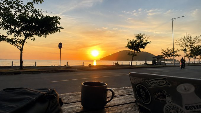 A scenic sunset view over the ocean from a wooden table with a coffee mug and a sticker-covered laptop in the foreground, showing a person on a motorbike on the coastal road.