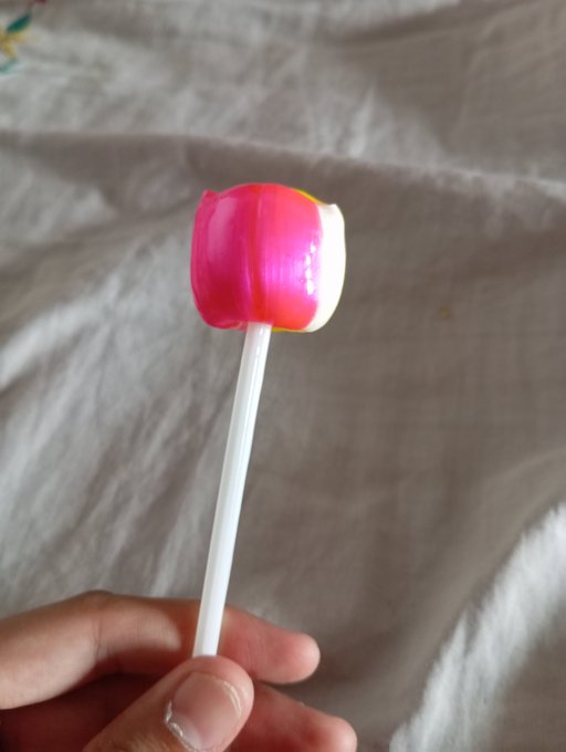 A hand holding a small pink and white mushroom-shaped lollipop on a white stick against a wrinkled white fabric background.