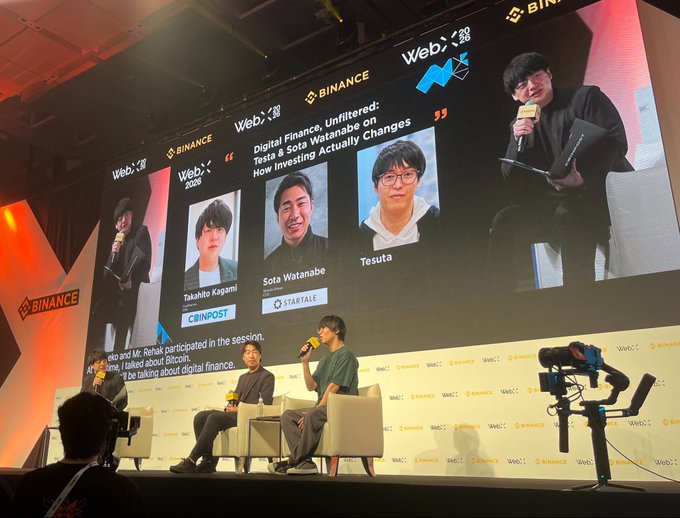 A panel discussion at WebX 2026, sponsored by Binance, features speakers including Takahashi Kogami (Coinpost) and Sota Watanabe (Startale) discussing "Digital Finance, Unfiltered: Tests & Sota Watanabe on How Investing Actually Changes," with a side screen showing Takahashi holding a microphone. This connects to the breaking news post where Testa reportedly mentioned being underwater on Bitcoin purchased from the previous event.