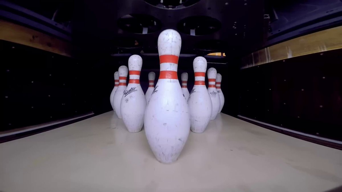 The image shows a row of white bowling pins lined up against each other with red stripes at the end. The pins are arranged in a straight line, creating an interesting visual effect. The background appears to be a dark room or alley, which contrasts with the bright and colorful arrangement of the bowling pins.