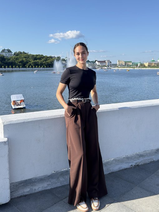 A young woman standing on a bridge over a lake, wearing a black t-shirt and brown satin wide-leg trousers, with a fountain and city buildings visible in the background.