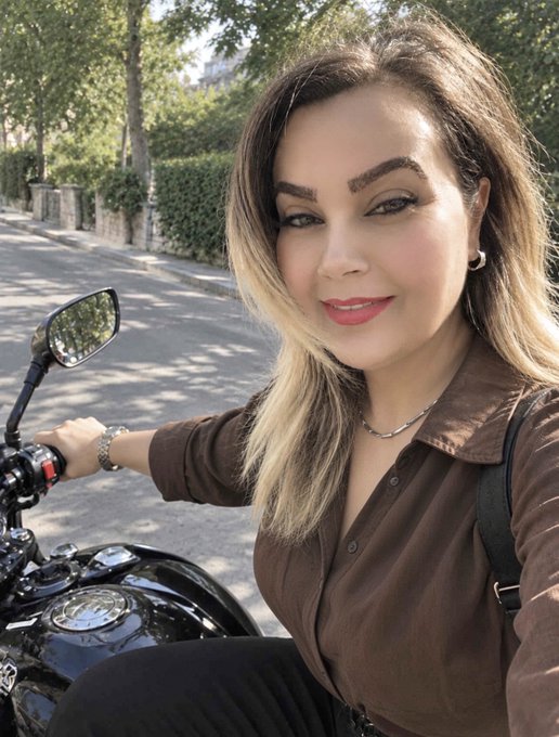 A woman with long blonde-highlighted hair, wearing a brown button-up shirt and silver necklace, sits on a black motorcycle while taking a selfie outdoors on a sunny day.