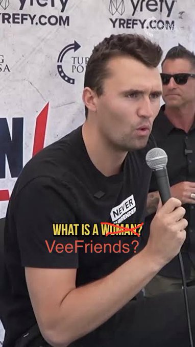 A man speaking into a microphone at a Yrefy event wearing a 'Never' t-shirt, with overlaid text reading 'WHAT IS A WOMAN?' and 'VeeFriends?'.