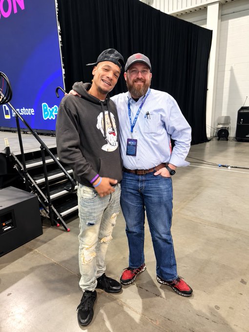 Two men posing together at what appears to be an event, with one wearing a grey graphic hoodie and backwards cap, and the other in a light blue shirt with a lanyard and red sneakers.