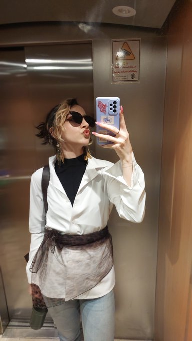 A woman with short hair takes a mirror selfie in an elevator wearing sunglasses, a white shirt over a black top, and a sheer brown sash, holding a phone with a purple case.