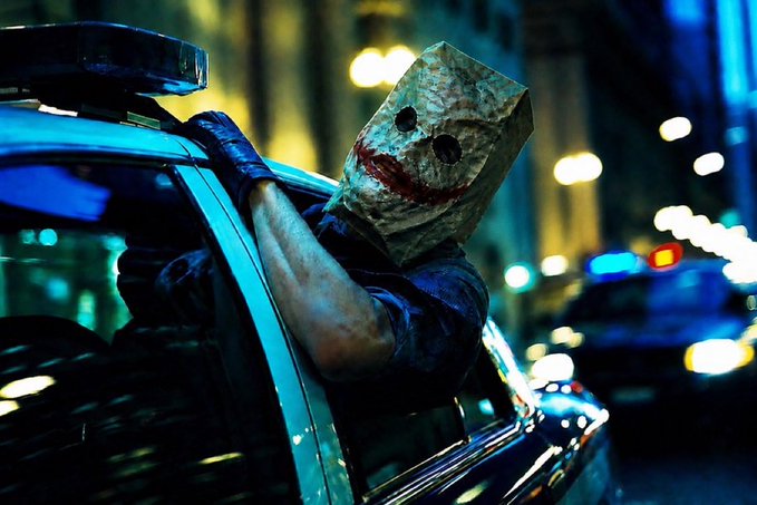 A figure wearing a paper bag mask with a sinister painted smile leans out of a police car window at night, with a blurred cityscape and another police cruiser with flashing lights in the background. The unsettling, chaotic imagery of a masked menace escaping police pursuit humorously mirrors the post's message of "zcat" violently dethroning "fartcoin" in a sudden "vibe shift."