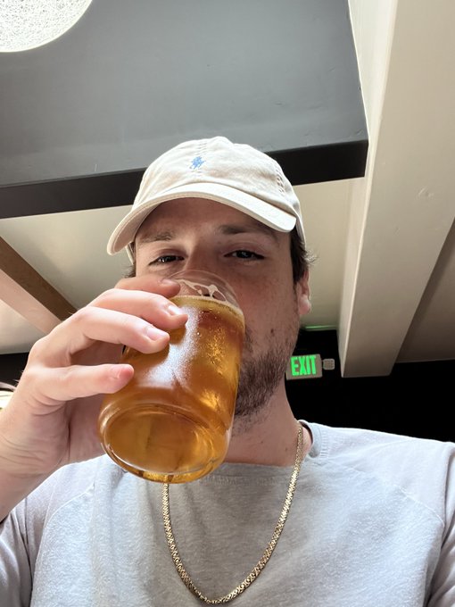 A man wearing a tan baseball cap and a gold chain takes a selfie, drinking from a plastic cup of beer.