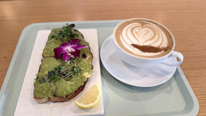 An overhead shot of a light green tray holding avocado toast garnished with microgreens and a purple orchid flower, alongside a white cup of latte art coffee (with a heart and cocoa powder design) and a lemon wedge.