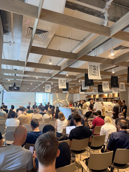 An audience attending a presentation in a modern room with exposed ceiling beams, where banners reading 'STABLE' and 'ASK ME ABOUT STABLE COINS' hang from the ceiling.
