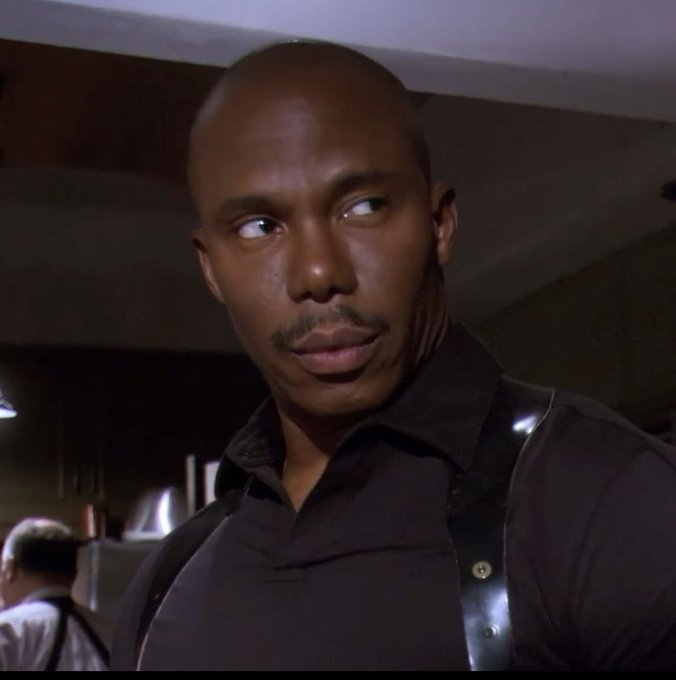 A man with a shaved head and a thin mustache looks off to the side with a suspicious, side-eyed expression, his brow slightly raised as if cautiously assessing something untrustworthy. His skeptical gaze perfectly captures the feeling of being wary of sudden, unexpected good fortune, much like a crypto investor suspicious of three consecutive green days in the market.