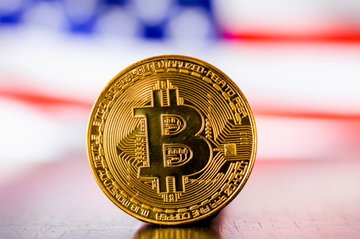 A golden physical Bitcoin token sits prominently in focus against a blurred American flag backdrop, visually symbolizing the intersection of cryptocurrency and U.S. legislative action as the Senate negotiates the #Bitcoin Clarity Act in Washington.