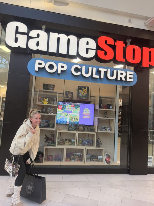 A woman poses in front of a GameStop store at the Mall of America, which features a large "GameStop POP CULTURE" sign above a display window filled with video games, collectibles, and a TV showing gameplay.