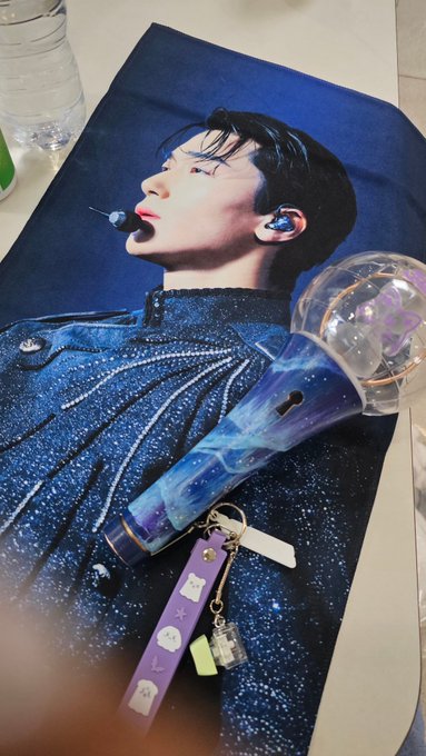 A young man with closed eyes lies on a dark pillow wearing an earpiece, with a sparkling blue lightstick and purple charms resting beside him, suggesting a K-pop fan photo.