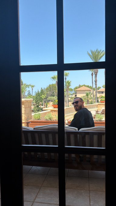 A man in sunglasses sits on an outdoor patio lounge chair, framed by a window looking out toward palm trees and resort-style buildings, capturing a relaxed morning vibe that fits the "Gm friends" greeting.