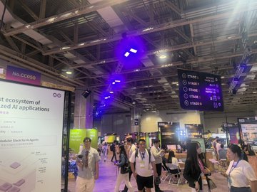 The image depicts a large convention center filled with people, many of whom are carrying backpacks. The scene appears to be set for an AI expo or event related to the application of AI systems. The blue lights above the crowd create an interesting visual effect, enhancing the atmosphere and drawing attention to the area. Various AI-related items can be seen throughout the venue, including a sign advertising "Web2+3板块比去年有了很大的扩张". The focus on this event highlights its importance in showcasing cutting-edge technologies and advancements within the field of AI.
