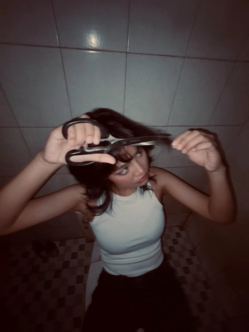 A high-angle photo of a young woman in a white tank top cutting her own dark hair with scissors in a dimly lit bathroom with tiled walls.