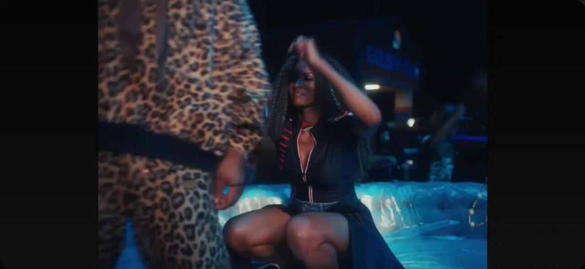 The image features a woman sitting in a pool, wearing a leopard print dress. The background of the image shows a man dressed as a leopard cat, possibly dancing or posing for a photo with the woman. There are also other people present in the scene, but they do not seem to be the main focus of the image.