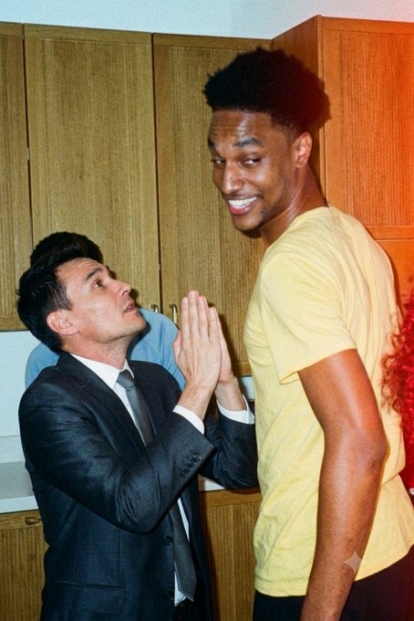 A photo showing a humorous height comparison between two men—a shorter man in a suit looking up with clasped hands, and a much taller, smiling man in a yellow t-shirt.