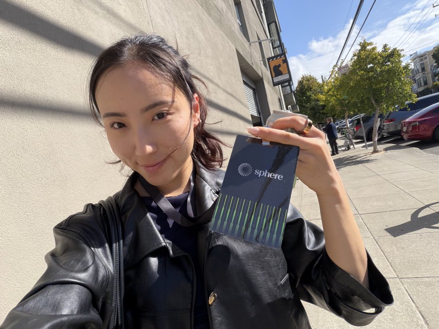 Selfie of a young woman in a black leather jacket standing on a sunny San Francisco sidewalk, holding up a 'Sphere' event badge.