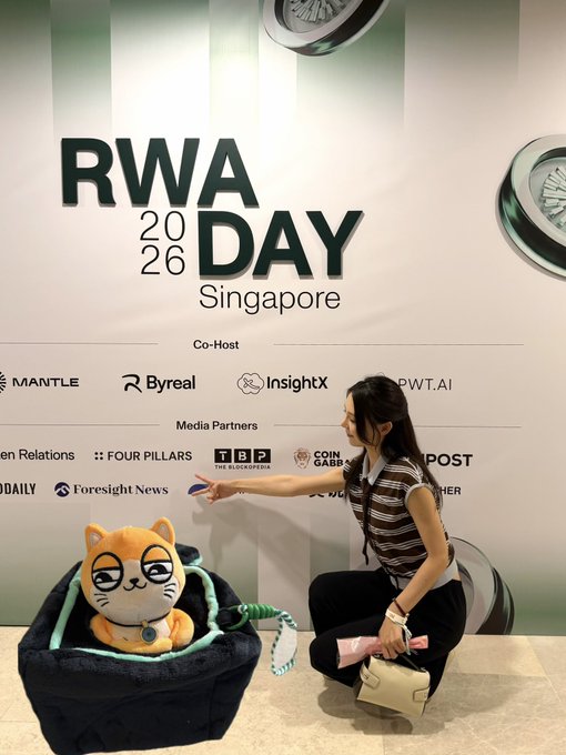 A woman in a brown striped shirt crouches next to a plush cat mascot at the 'RWA Day 2026 Singapore' event, with co-hosts Mantle, Byreal, InsightX, and PWT.AI listed on the backdrop.
