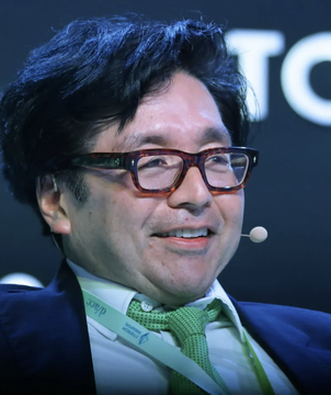 The image features a man wearing glasses with brown leather frames and a green necktie, sitting behind a desk. He has a happy expression on his face as he looks directly at the camera. The background of the photo contains text that reads "BITMINE FILES FOR PREFERRED STOCK OFFERING WITH A 9.5% YIELD Bitmine is raising fresh capital just weeks after its largest ETH purchase of 2026, edging closer to its goal of owning 5% of Ethereum'", suggesting a connection between the man's attire and the cryptocurrency market.