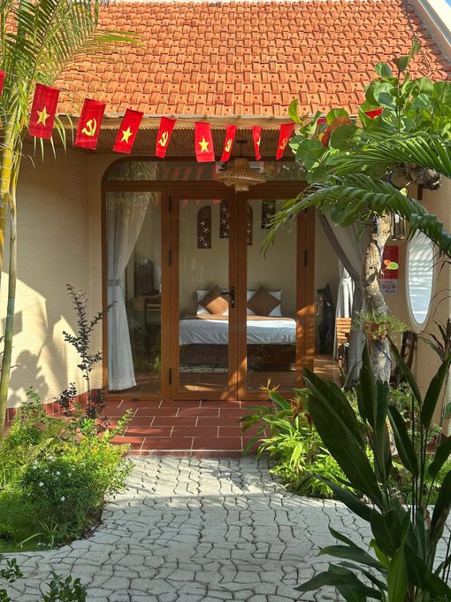 The image shows an outdoor area with a house, featuring red roof tiles that give it a Vietnamese-inspired look. A bed can be seen inside the house, suggesting that this space may serve as both a living room and bedroom. There are also flags hanging from the roof of the house, adding to its unique character. In front of the house, there is a garden with various plants and trees, creating a peaceful atmosphere. The post accompanying the image provides information about the house's features and possibly some cultural context related to Vietnam.